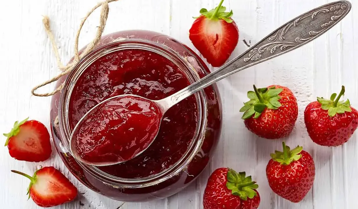 Glas mit Erdbeermarmelade, gesüßt mit Xylit, daneben frische Erdbeeren und ein Löffel Marmelade.