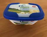 Eine Packung "Biofree Laktosefrei Teebutter" mit blauem Deckel steht auf einem Holztisch. Die Verpackung zeigt eine Sommerwiese mit Blumen und Butterflocken.