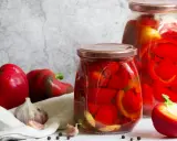 Frische rote Paprika, Knoblauch und Pfefferkörner neben zwei Gläsern mit eingelegter Paprika. Perfekt zum Paprika einkochen mit Weck-Gläsern.