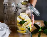 Einmachgläser werden mit marokkanischen Salzzitronen gefüllt. Eine Hand gießt mit einer kleinen Schöpfkelle eine Flüssigkeit über die Zitronen.