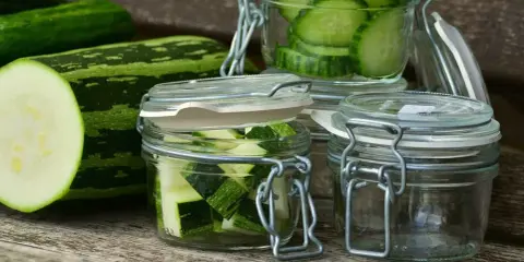 Zucchini-Scheiben und -Würfel in Gläsern, bereit zum einkochen im Dampfgarer. Frische Zucchini liegt daneben.