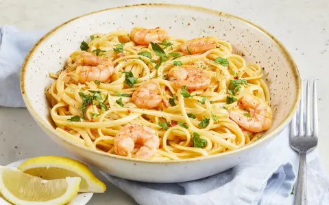 Eine Schüssel mit Spaghetti mit Garnelen, garniert mit Petersilie und Chiliflocken. Dazu Zitronenspalten und eine Gabel.