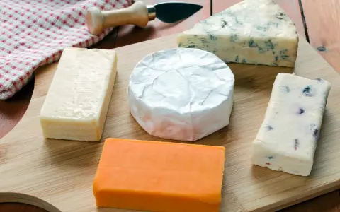 Verschiedene Käsesorten auf einem Holzbrett. Warum ist Cheddar orange? Die natürliche Farbe kommt von Beta-Carotin, das Kühe über Gras aufnehmen.