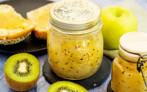 Ein Glas mit leuchtend gelber kiwi-marmelade mit schale, daneben frische Kiwischeiben und geröstetes Brot.