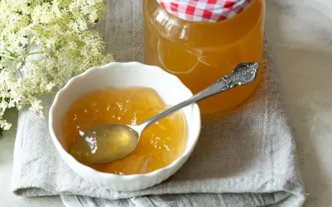 Ein Löffel voll goldgelbem Holunder Apfel Gelee in einer Schale, daneben ein Glas mit dem Gelee und Holunderblüten.