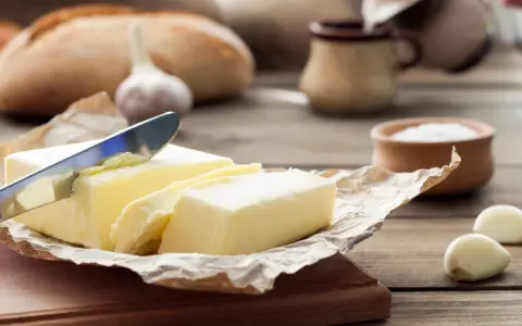 Ein Messer schneidet ein Stück von einem Block welcher Butter. Daneben liegen Knoblauch und ein Brot.