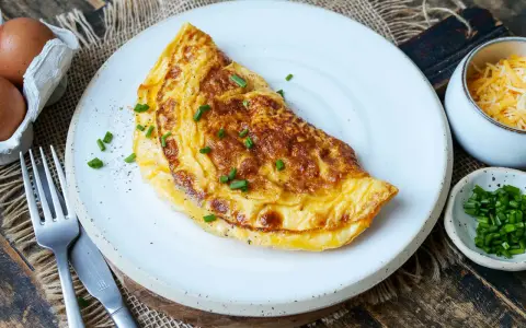 Goldbraunes Omelett mit Käse und Schnittlauch, perfekt für ein schnelles Omelette Rezept. Daneben: Eier, geriebener Käse und frischer Schnittlauch.