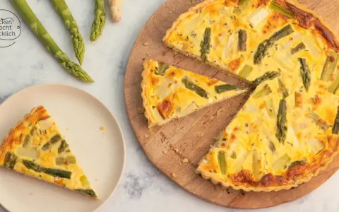 Herzhafte Spargel Quiche mit frischem Spargel, goldbraun gebacken. Ein Stück auf einem Teller, der Rest auf einem Holzbrett.