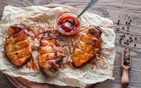 Saftiges Grillfleisch zart machen: Drei gegrillte Hähnchenbrustfilets mit Sauce auf Backpapier, daneben eine Schale Ketchup und eine Holzschaufel mit Pfefferkörnern.