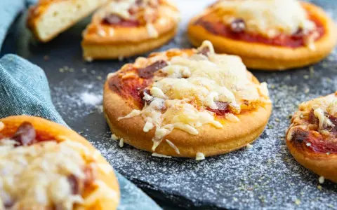 Mehrere mini pizza mit geschmolzenem Käse und Tomatensauce auf einem dunklen Untergrund, bestreut mit etwas Mehl.