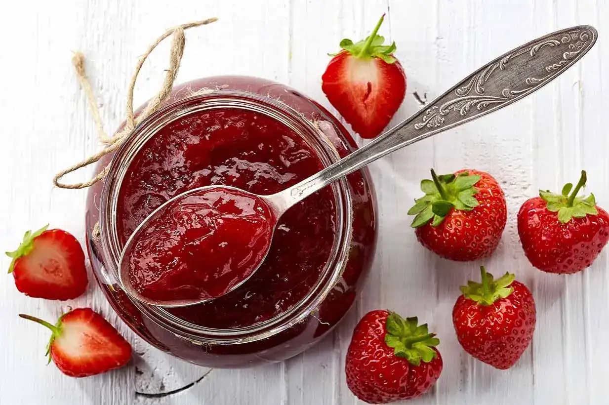 Glas mit Erdbeermarmelade, gesüßt mit Xylit. Frische Erdbeeren und ein Löffel Marmelade liegen daneben.