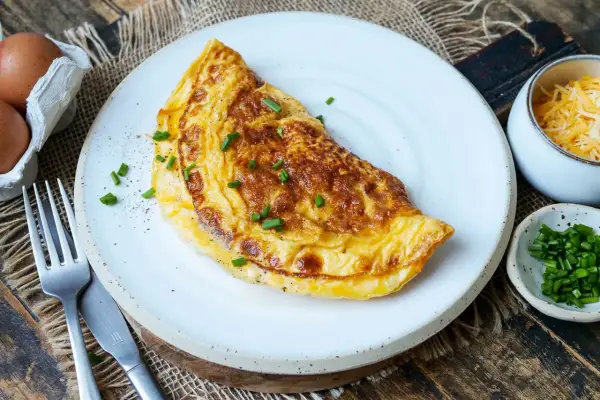 Goldbraunes Omelett mit Käse und Schnittlauch, perfekt für ein schnelles Omelette Rezept. Daneben: Eier, geriebener Käse und frischer Schnittlauch.