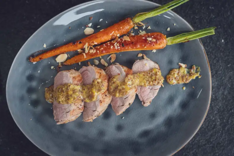 Zartes schweinefilet grillen, serviert mit karamellisierten Karotten und grobem Senf. Geröstete Mandelsplitter runden das Gericht ab.