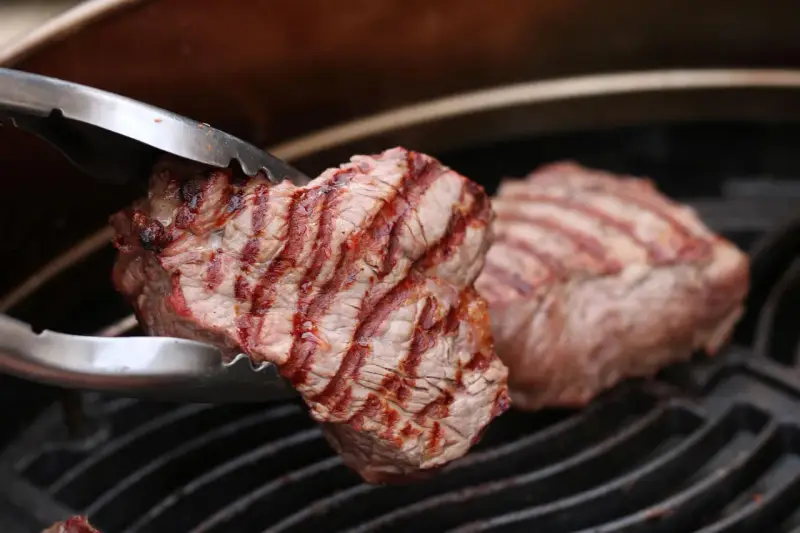 Zangen halten ein saftiges Stück Roastbeef, das auf einem Grill zubereitet wird.